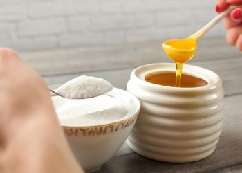 carboidrati semplici: una ciotola con zucchero bianco e una con del miele. Le mani di una donna tengono il cucchiaio dell'una e dell'altra