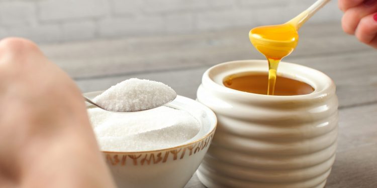 carboidrati semplici: una ciotola con zucchero bianco e una con del miele. Le mani di una donna tengono il cucchiaio dell'una e dell'altra