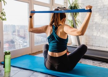 Donna a casa di schiena seduta su un tappeto blu vicino alla finestra allena il muscolo deltoide con un elastico fitness. Accanto a lei una borraccia verde.