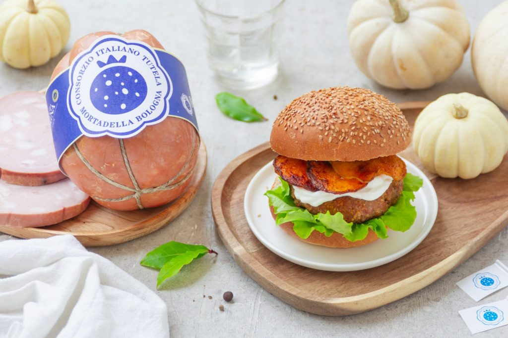 Burger con mortadella posizionato su un piatto di legno e accompagnato da elementi di decorazione
