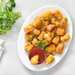Nuggets di pollo al forno: panatura dorata e super croccante 1 In primo piano su un piatto ovale bianco ci sono dei nuggets di pollo con una ciotolina di salsa ketchup.