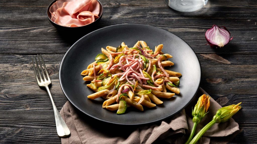 su tavolo di legno scuro, piatto nero con dentro pasta con mortadella e zucchine, accanto un piattino con mortadella e in basso a destra 2 fiori di zucchina accanto alla forchetta