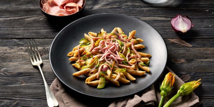 su tavolo di legno scuro, piatto nero con dentro pasta con mortadella e zucchine, accanto un piattino con mortadella e in basso a destra 2 fiori di zucchina accanto alla forchetta