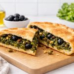In primo piano su un tagliere di legno chiaro una pizza di scarola con olive e capperi e acciughe. Sullo sfondo ci sono un'oliera, una ciotola bianca con delle olive nere e la scarola.