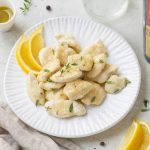 Pollo al limone in padella, un secondo leggero e saporito 2 In primo piano su uno sfondo chiaro c'è un piatto con del pollo al limone, decorato con delle fettine di limone. A destra si trova una bottiglia di olio di Roma IGP.