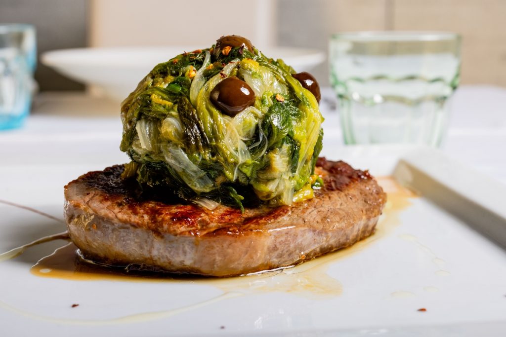 su tavolo bianco: una fetta di carne di manzo con scarola in padella sopra a forme di palle con olive nere di Gaeta