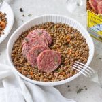 su tavolo con tovaglia bianca, insalatiera di ceramica bianca con cotechino e lenticchie , sulla destra piatto pieno della pietanza con forchetta e dietro confezione della scatola del cotechino Modena IGP