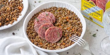 su tavolo con tovaglia bianca, insalatiera di ceramica bianca con cotechino e lenticchie , sulla destra piatto pieno della pietanza con forchetta e dietro confezione della scatola del cotechino Modena IGP