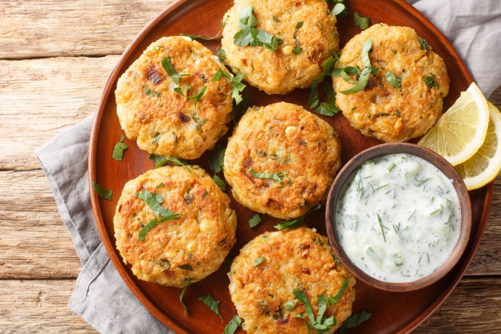 Hamburger di ceci: una ricetta vegetariana semplice 3 su tavolo di legno, canovaccio e sopra un piatto marrone con hamburger di ceci e prezzemolo e un contenitore con salsa allo yogurt e 2 fette di limone