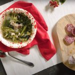 su tavolo nero tagliere bianco con un contenitore con minestra maritata con salamini sulla destra; tagliere di legno con un salame cacciatore e sulla sinistra 2 rami di legno con fiocco rosso
