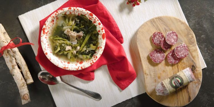 su tavolo nero tagliere bianco con un contenitore con minestra maritata con salamini sulla destra; tagliere di legno con un salame cacciatore e sulla sinistra 2 rami di legno con fiocco rosso