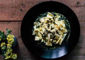 su tavolo di legno, piatto nero con pasta con spinaci freschi. A sinistra una pianta con fiori