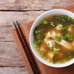 Zuppa di miso: la ricetta da fare a casa con step by step fotografico 1 In primo piano una zuppa di miso con cubetti di tofu, alghe wakame e cipollotto verde in una ciotola bianca su tavolo in legno con bacchette.