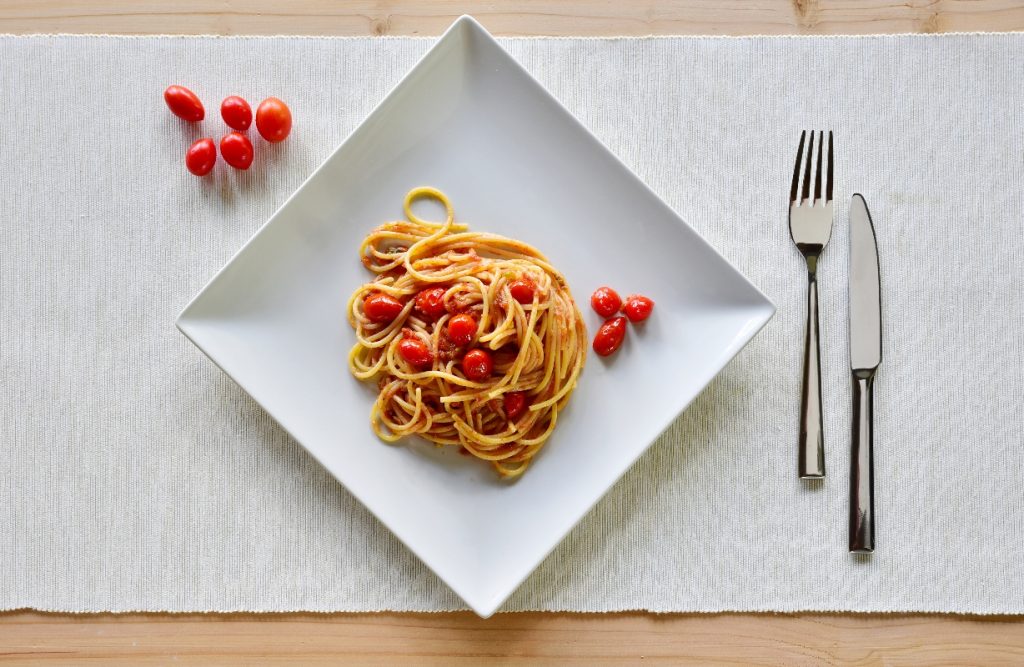 Pasta con pomodorini: un primo piatto veloce e gustoso 4 su tavolo di legno sottopiatto bianco di stoffa con un un piatto quadrato pieno di pasta con pomodorini freschi. Accanto al piatto 5 pomodorini e a destra forchette e coltello