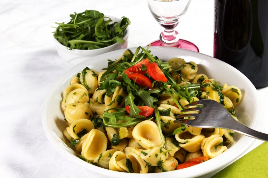 Pasta con rucola e pomodoro: un primo piatto sano e veloce 3 su tavolo con tovaglia bianca, un piatto bianco con pasta con rucola e pomodoro (orecchiette), vicino un bicchiere d'acqua e un contenitore di rucola