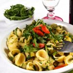 su tavolo con tovaglia bianca, un piatto bianco con pasta con rucola e pomodoro (orecchiette), vicino un bicchiere d'acqua e un contenitore di rucola