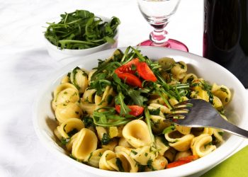 su tavolo con tovaglia bianca, un piatto bianco con pasta con rucola e pomodoro (orecchiette), vicino un bicchiere d'acqua e un contenitore di rucola