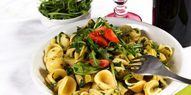 su tavolo con tovaglia bianca, un piatto bianco con pasta con rucola e pomodoro (orecchiette), vicino un bicchiere d'acqua e un contenitore di rucola