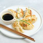 In primo piano su un tavolo di marmo bianco c'è un piatto bianco fondo con dei ravioli cinesi. Accanto una piccola ciotola con la salsa di soia e due bacchette di legno.