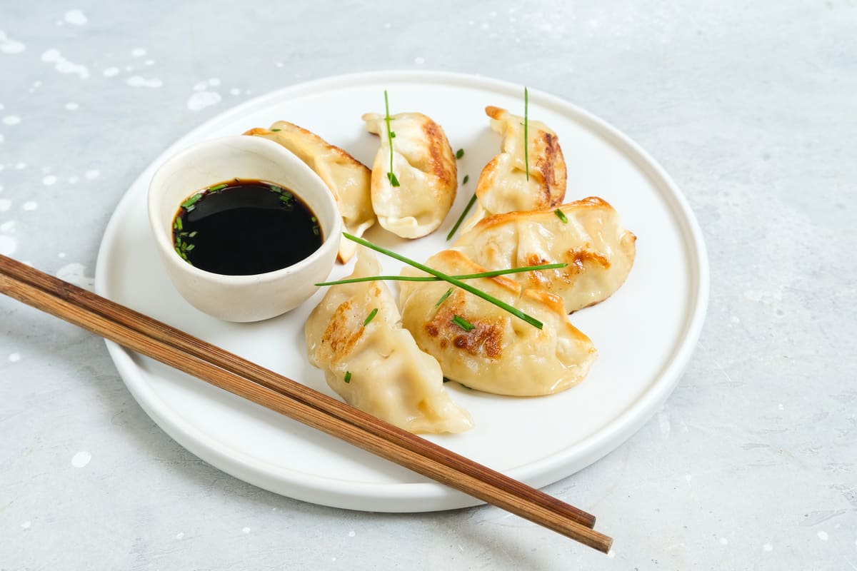 In primo piano su un tavolo di marmo bianco c'è un piatto bianco fondo con dei ravioli cinesi. Accanto una piccola ciotola con la salsa di soia e due bacchette di legno.