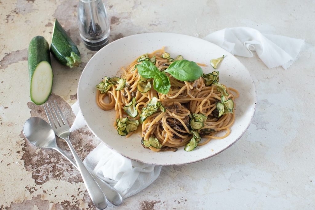 Spaghetti alla Nerano: un primo piatto vegetariano semplice e genuino 3 su tavolo di marmo, piatto fondo con spaghetti alla Nerano con zucchine e basilico, sulla sinistra cucchiaio e forchetta e dietro il piatto una zucchina tagliata in 2 e un contenitore pepe