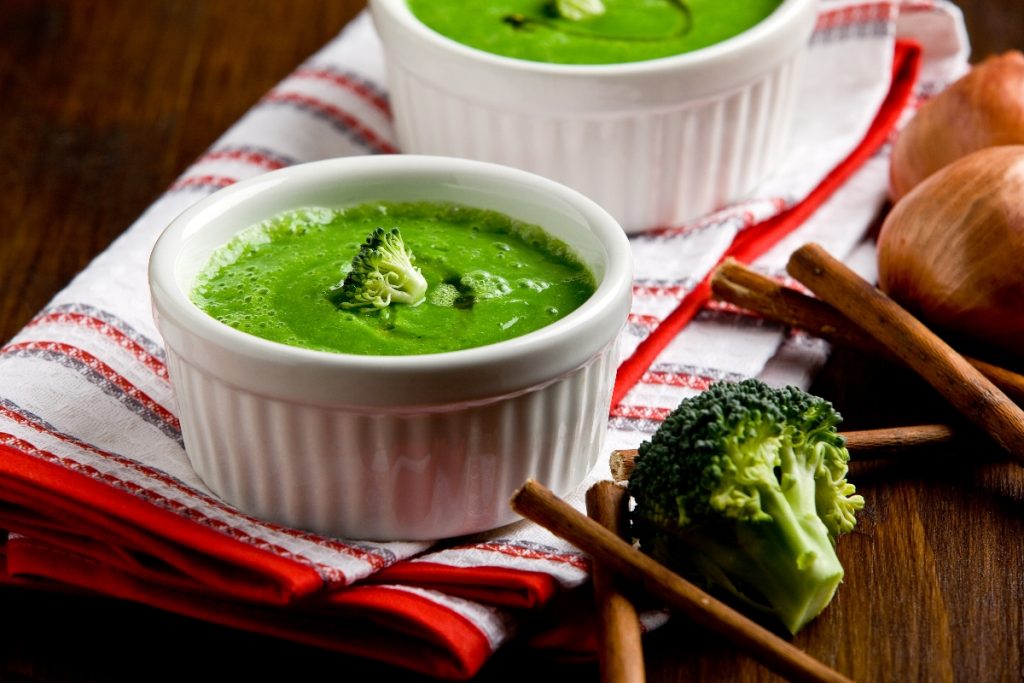 Vellutata di broccoli: una ricetta cremosa e dietetica 1 su tavolo di legno, canovaccio bianco e rosso piegato con 2 ciotole piccole con vellutata di broccoli con bacchette cinesi e accanto un pezzo di broccolo