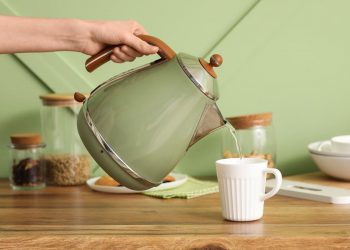 tavolo apparecchiato per fare uno spuntino o una colazione, in primo piano una mano che tiene un bollitore elettrico verde e versa dell'acqua calda in una tazza bianca