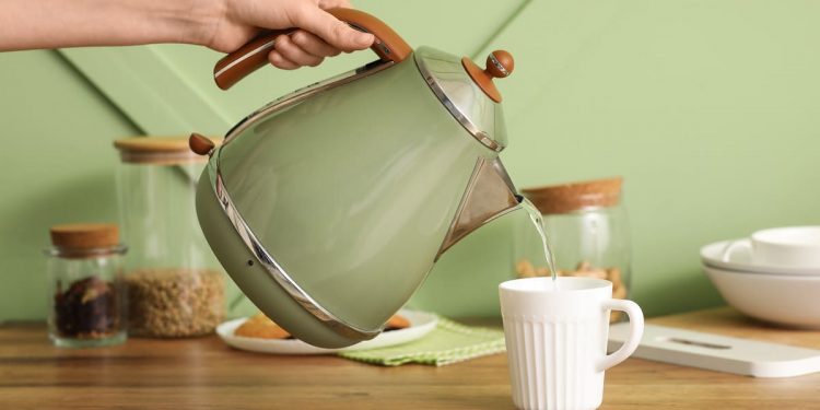 tavolo apparecchiato per fare uno spuntino o una colazione, in primo piano una mano che tiene un bollitore elettrico verde e versa dell'acqua calda in una tazza bianca