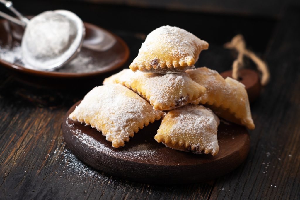 su tavolo di legno, tagliere con bugie ripiene di cioccolato, in fondo zucchero velato con un setaccio