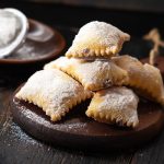 su tavolo di legno, tagliere con bugie ripiene di cioccolato, in fondo zucchero velato con un setaccio