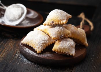 su tavolo di legno, tagliere con bugie ripiene di cioccolato, in fondo zucchero velato con un setaccio