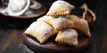 su tavolo di legno, tagliere con bugie ripiene di cioccolato, in fondo zucchero velato con un setaccio