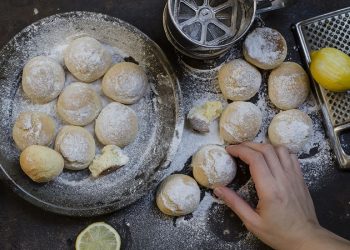 su tavolo nero, piatto con le castagnole al forno, una mano le passa nello zucchero velato , accanto una grattugia con limone e in alto un setaccio