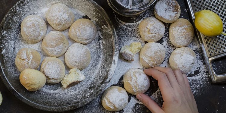 su tavolo nero, piatto con le castagnole al forno, una mano le passa nello zucchero velato , accanto una grattugia con limone e in alto un setaccio