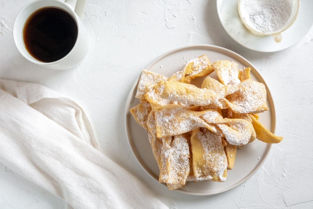 tavolo bianco da colazione con chiacchiere senza glutine in un piatto bianco, sulla sinistra una tazza di caffè, accanto un tovagliolo bianco