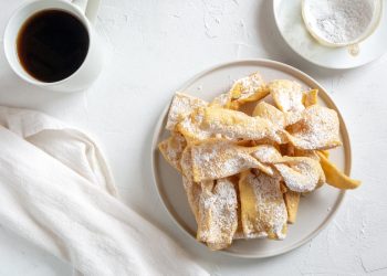 tavolo bianco da colazione con chiacchiere senza glutine in un piatto bianco, sulla sinistra una tazza di caffè, accanto un tovagliolo bianco