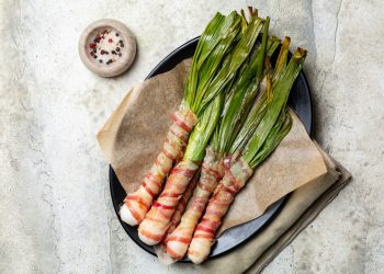 su tavolo bianco piatto nero con carta da forno, cipollotti al forno con speck attorno al gambo, vicino ciottola con pepe nero