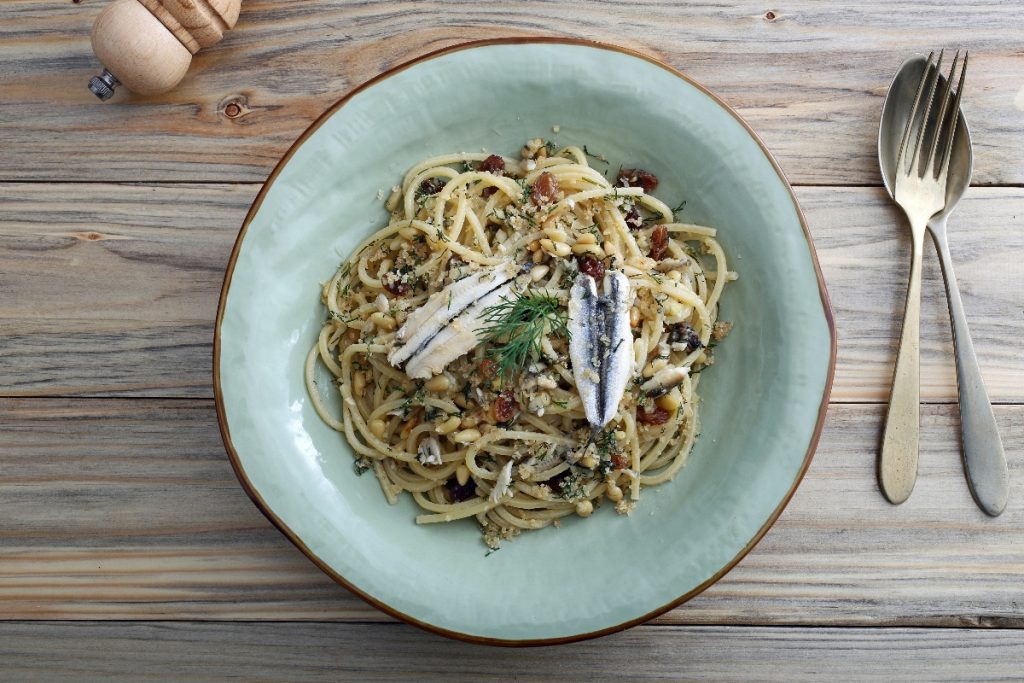Pasta con le alici fresche: un primo piatto sano e saporito 2 su tavolo di legno piatto fondo azzurro con pasta con le alici; sulla destra cucchiaio e forchetta