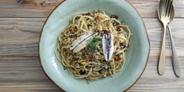 su tavolo di legno piatto fondo azzurro con pasta con le alici; sulla destra cucchiaio e forchetta