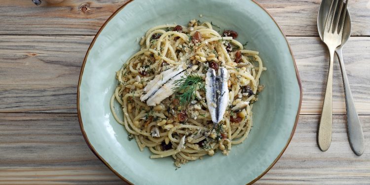 su tavolo di legno piatto fondo azzurro con pasta con le alici; sulla destra cucchiaio e forchetta