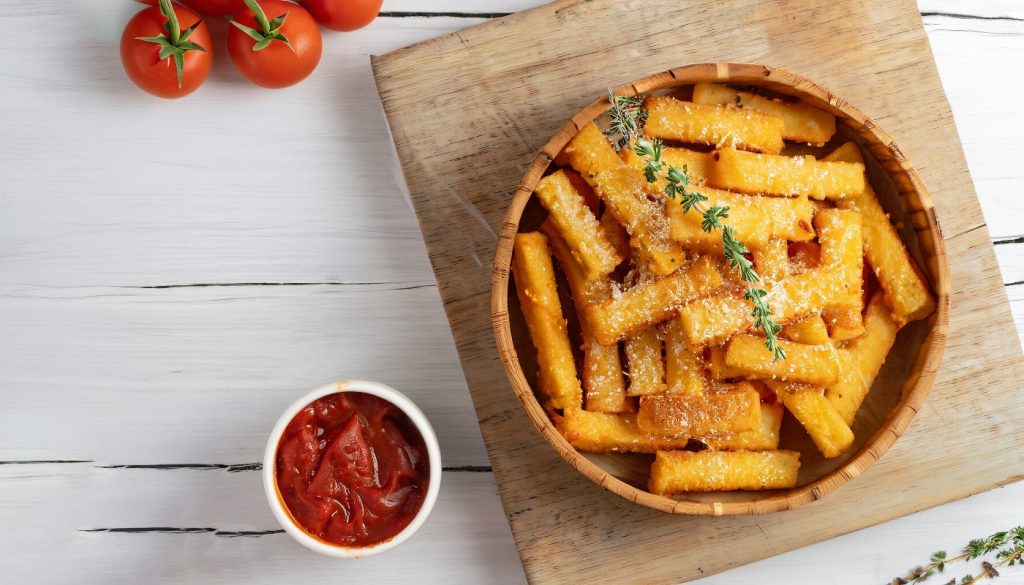 su tavolo bianco tagliere di legno con ciotola con la polenta al forno e parmigiano grattugiato e erbe, accanto piccola ciotola con salsa di pomodoro e dietro pomodorini