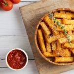 su tavolo bianco tagliere di legno con ciotola con la polenta al forno e parmigiano grattugiato e erbe, accanto piccola ciotola con salsa di pomodoro e dietro pomodorini