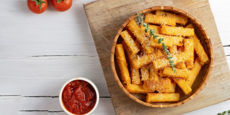 su tavolo bianco tagliere di legno con ciotola con la polenta al forno e parmigiano grattugiato e erbe, accanto piccola ciotola con salsa di pomodoro e dietro pomodorini