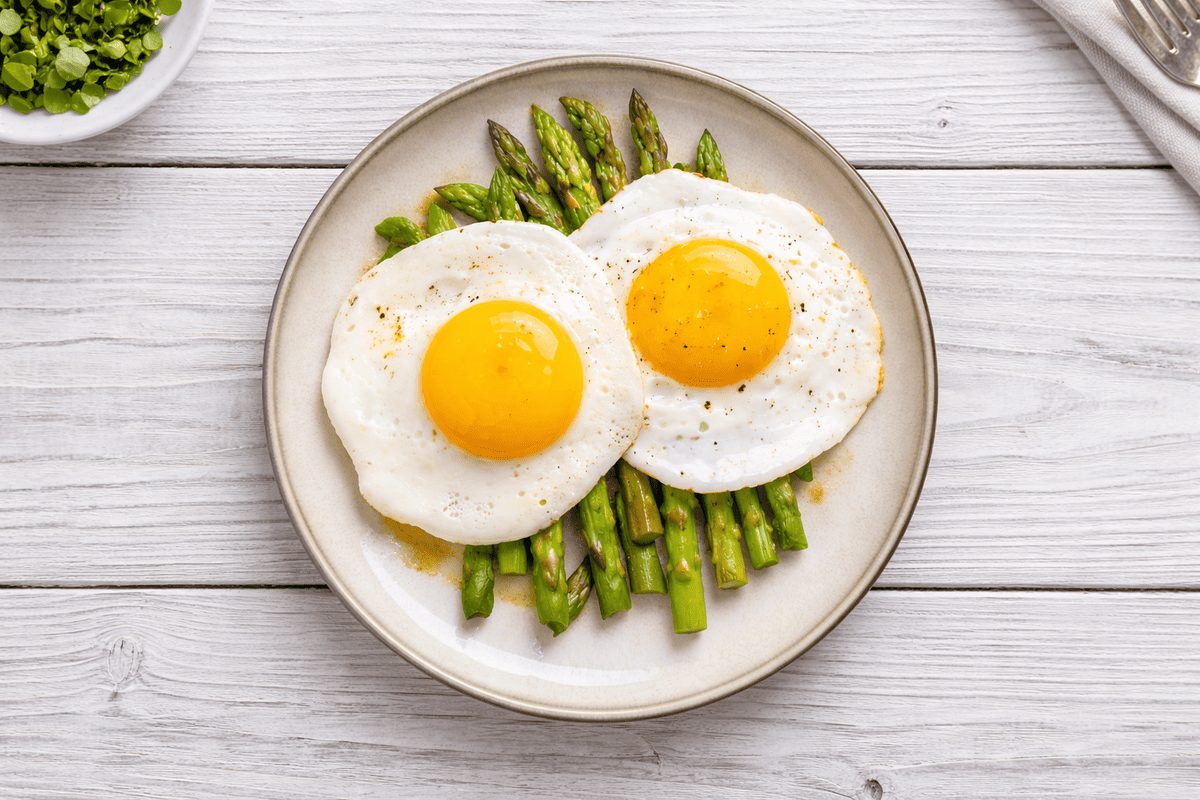 In primo piano su un tavolo di legno chiaro c'è un piatto fondo con degli asparagi alla milanese.