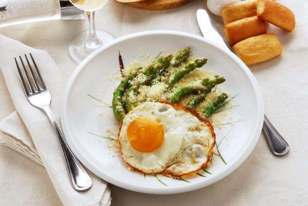 Asparagi alla milanese: un secondo piatto delizioso 2 su una tavolo apparecchiata, piatto bianco con asparagi alla milanese con asparagi e uova con forchetta e coltello con tovaglioli bianchi , un calice di vino e panini