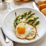su una tavolo apparecchiata, piatto bianco con asparagi alla milanese con asparagi e uova con forchetta e coltello con tovaglioli bianchi , un calice di vino e panini