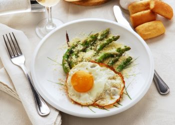 su una tavolo apparecchiata, piatto bianco con asparagi alla milanese con asparagi e uova con forchetta e coltello con tovaglioli bianchi , un calice di vino e panini