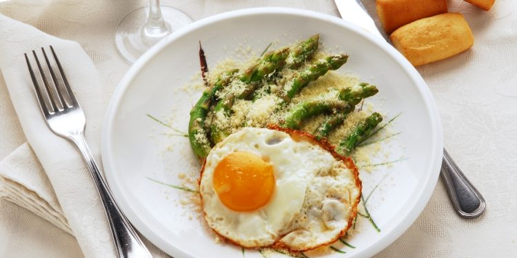 su una tavolo apparecchiata, piatto bianco con asparagi alla milanese con asparagi e uova con forchetta e coltello con tovaglioli bianchi , un calice di vino e panini