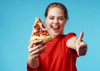 su sfondo blu chiaro donna con maglietta rossa felicissima che fa l'occhietto e il pollice in alto perché segue il cheat day e nell'altra mano tiene un pezzo di pizza con salame