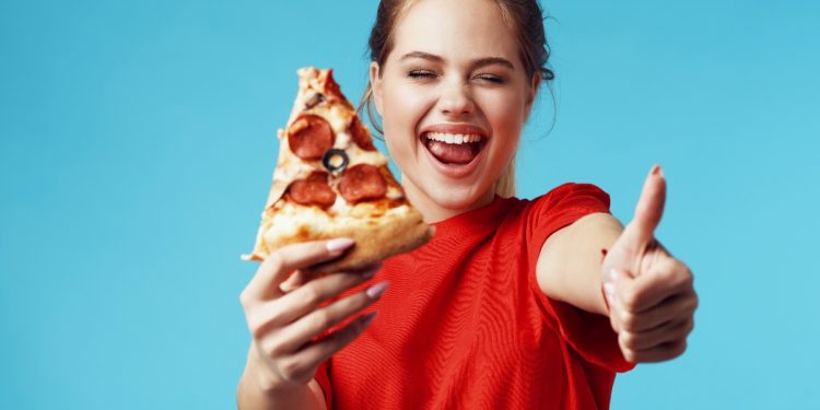 su sfondo blu chiaro donna con maglietta rossa felicissima che fa l'occhietto e il pollice in alto perché segue il cheat day e nell'altra mano tiene un pezzo di pizza con salame